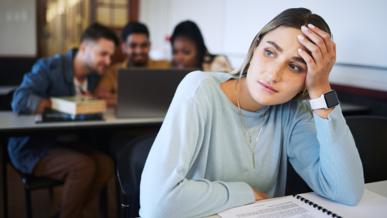 Social-Anxiety-Do-you-experience-these-symptoms Young woman in a room sitting alone with an anxious expression while a group of 3 people are talking and working together behind her | Social Anxiety Do you experience these symptoms | Mindful Health Solutions
