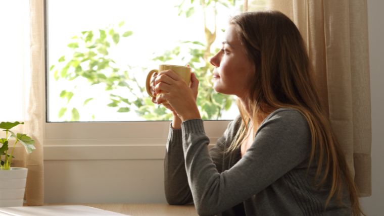 TMS-Side-Effects-Important-Facts-You-Should-Know Woman drinking a warm beverage while looking out the window on a sunnday day | TMS Side Effects Important Facts You Should Know | Mindful Health Solutions