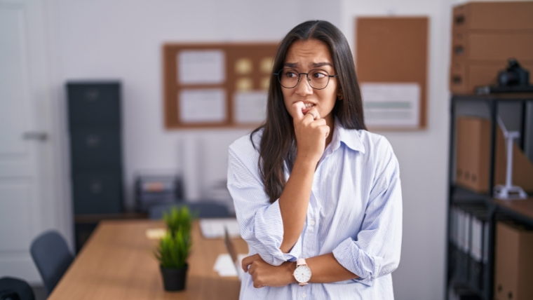 Identifying-Imposter-Syndrome-Uncover-6-Signs-and-Take-a-Self-Assessment Young woman of color at the office looking stressed and nervous with hands on mouth biting nails. | Identifying Imposter Syndrome Uncover 6 Signs and Take a Self-Assessment | Mindful Health Solutions