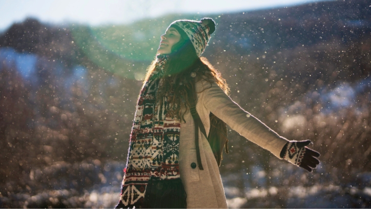 Boost-Your-Well-being-with-9-Mindfulness-Practices-This-Holiday-Season Woman standing outside in woods while it is snowing. Her arms are out and her head is titled up, enjoying the snow fall. | Boost Your Well-being with 9 Mindfulness Practices This Holiday Season | Mindful Health Solutions