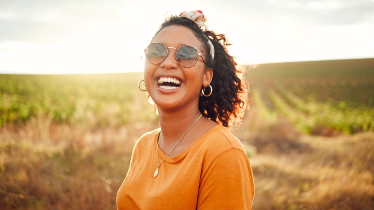 The-Rise-of-Esketamine-7-Reasons-Why-Its-Becoming-an-Antidepressant-Alternative A woman of color outside in a field wearing an orange shirt and sunglasses smiling | The RIse of Esketamine: 7 Reasons Why It's Becoming an Antidepressant Alternative | Mindful Health Solutions