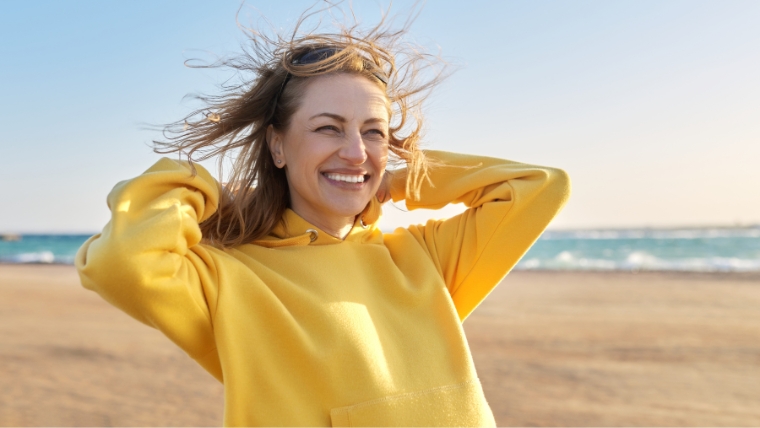 How To Say Goodbye to Depression FAST Introducing Accelerated TMS Caucasian woman on beach, hair being blown by wind, with a big smile on her face | How To Say Goodbye to Depression FAST Introducing Accelerated TMS | Mindful Health Solutions
