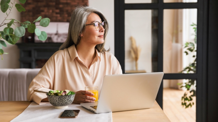TMS-vs.-Traditional-Pain-Management-Which-Is-Right-for-Your-Chronic-Pain Mature woman sitting at a table with a salad and computer, looking off in the distance thinking | TMS vs. Traditional Pain Management Which Is Right for Your Chronic Pain | Mindful Health Solutions