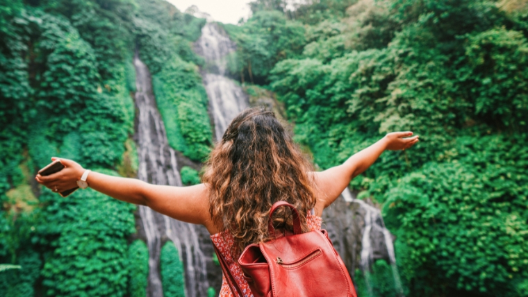 Say-Goodbye-to-Travel-Anxiety-8-Simple-Ways-To-Have-a-Stress-Free-Trip Woman facing a waterfall with a backpack on and her arms wide open | Say Goodbye to Travel Anxiety 8 Simple Ways To Have a Stress-Free Trip | Mindful Health Solutions