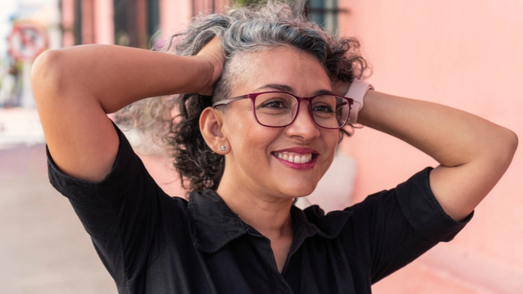 TMS Therapy Treatment for Chronic Pain Relief Is It Right for You Mature woman with glasses smiling in front a pink wall with her hands in her hair | TMS Therapy Treatment for Chronic Pain Relief: Is It Right for You? | Mindful Health Solutions